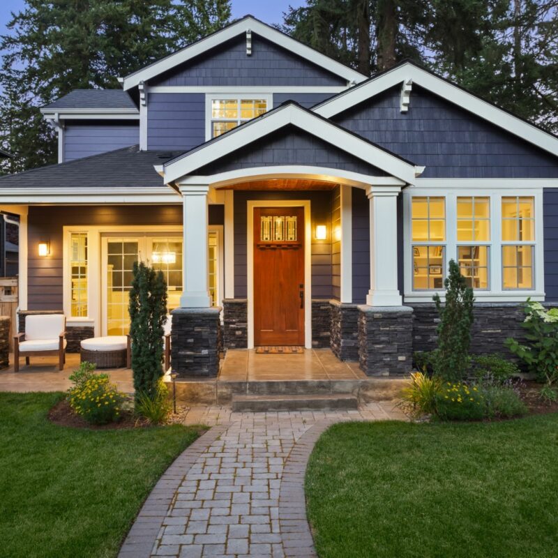 Modern two-story home with dark blue siding, white columns, and warm lighting illuminating the red front door and large windows at dusk