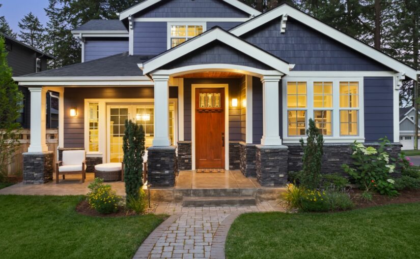 Modern two-story home with dark blue siding, white columns, and warm lighting illuminating the red front door and large windows at dusk