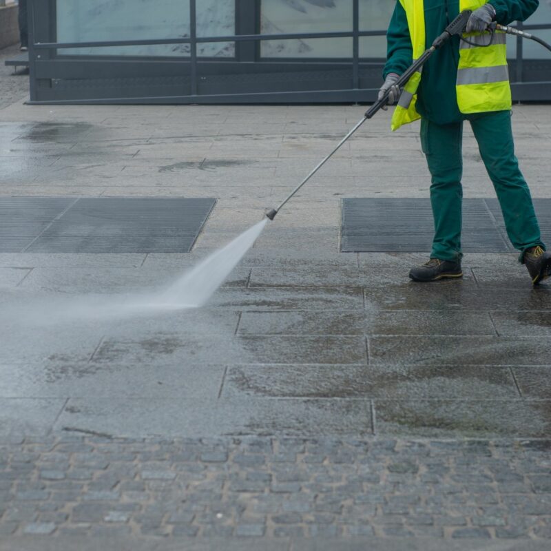 Worker in safety vest pressure washing urban pavement with high-powered water jet