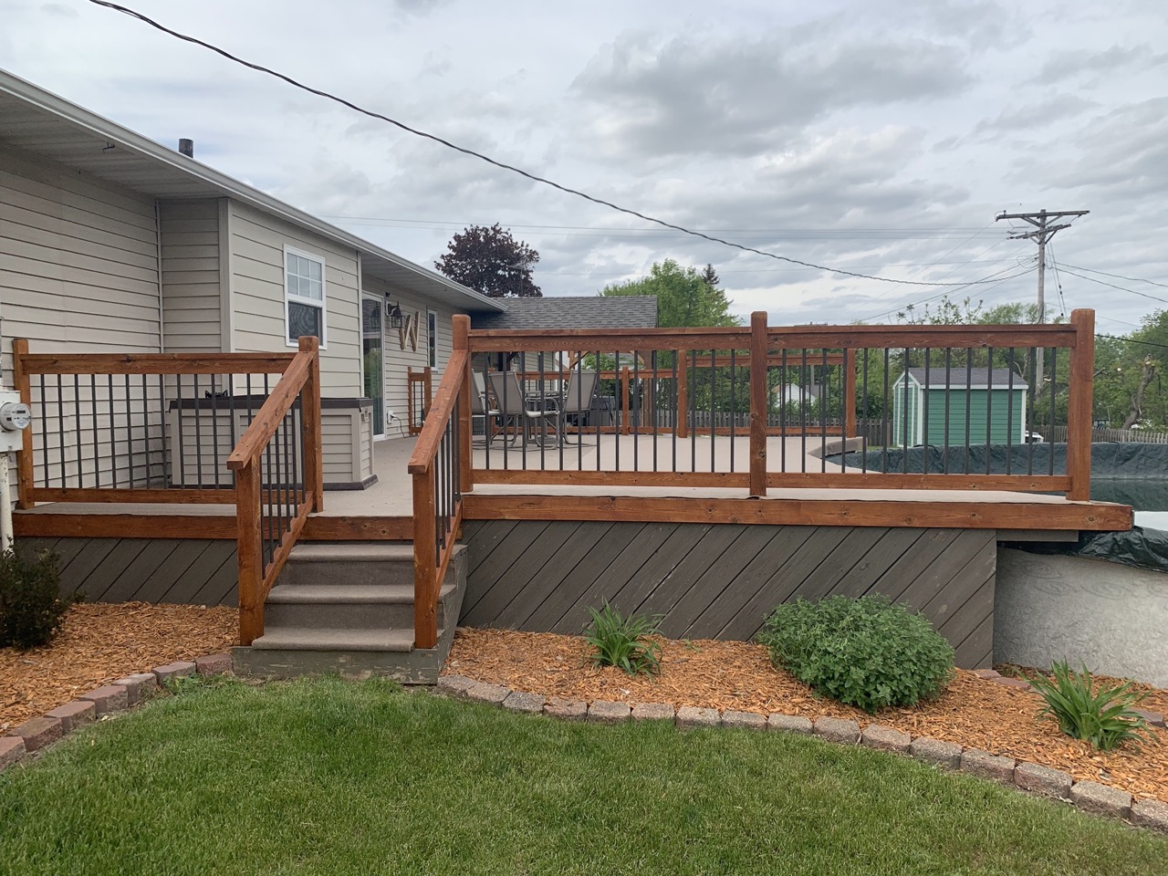 Two-story residential house in Fond du Lac with brown wooden deck railing and composite decking, featuring metal balusters and steps, surrounded by landscaped yard with mulch and shrubs