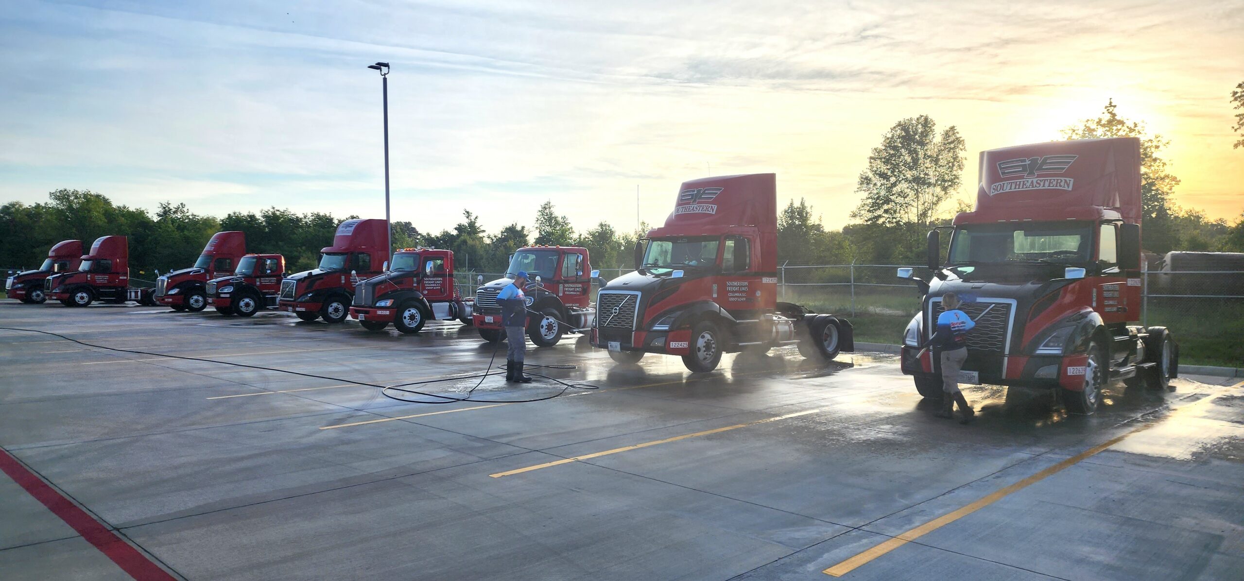 Fleet of red Southeastern semi trucks being pressure washed in commercial lot during sunset in Texarkana