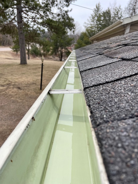 Clean green gutter system along dark asphalt roof shingles with forested landscape in background