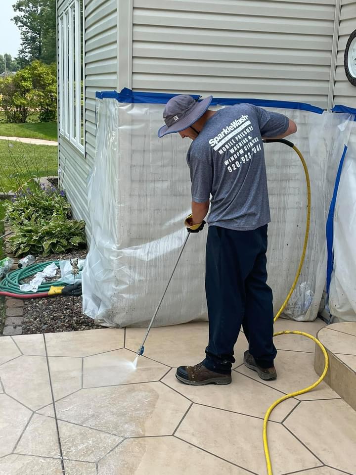 Worker pressure washing a concrete patio next to a mobile home with protective plastic sheeting in Fond du Lac, Wisconsin